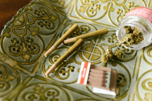 Marijuana spills out of a container as three joints and a pack of matches lay on a plate