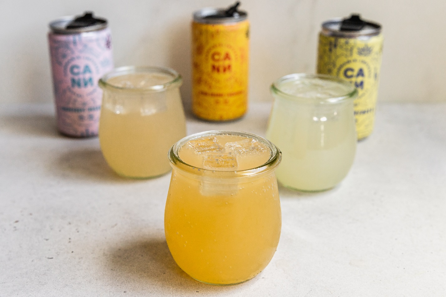 Three glasses with drinks in orange, yellow, and white sit in front of opened cans on a countertop