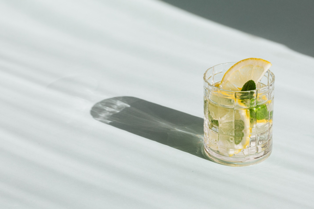 A glass with a drink, mint leaves, and lemon slices sits on a white surface, casting a dark shadow