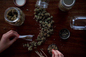 Hands are seen rolling a joint with cannabis scattered on a wooden surface