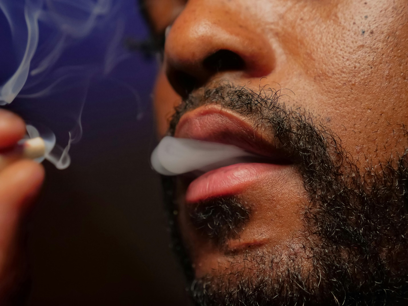 A close-up of a bearded man's nose and mouth as smoke exits a small gap between his lips, holding a joint between his thumb and forefinger amidst a cloud of smoke, epitomizing the enjoyment of cannabis pre-rolls.