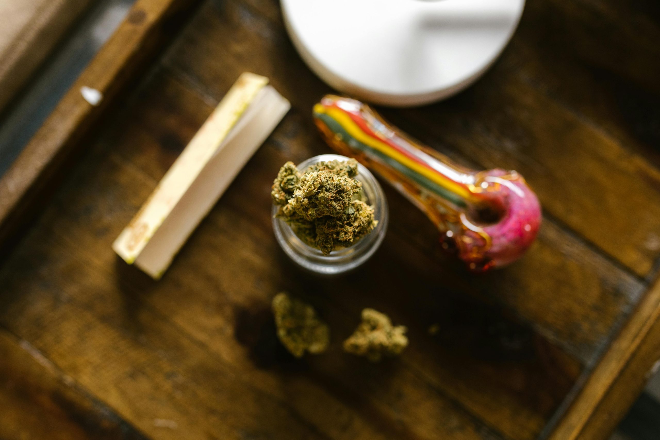 Cannabis buds in a jar with a colorful pipe and rolling papers on a wooden surface
