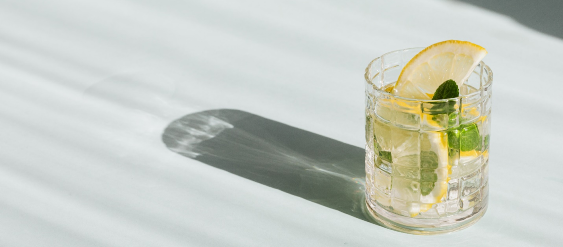 A glass with a drink, mint leaves, and lemon slices sits on a white surface, casting a dark shadow